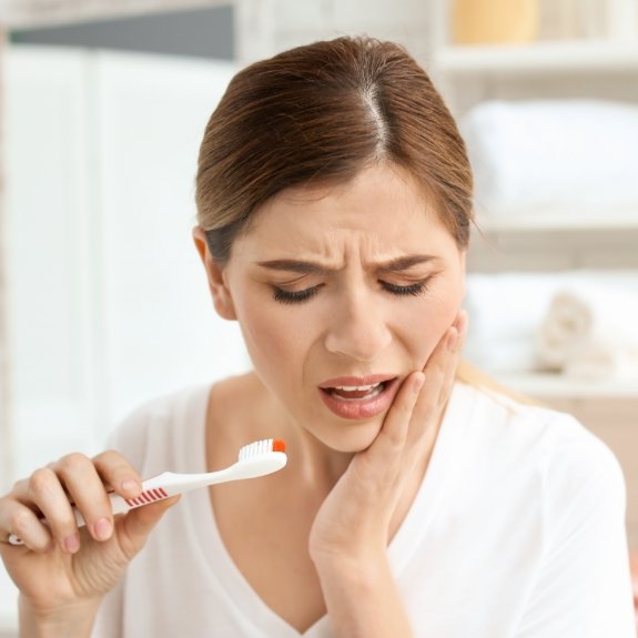 Woman holding her cheek in pain in one hand and her toothbrush in the other